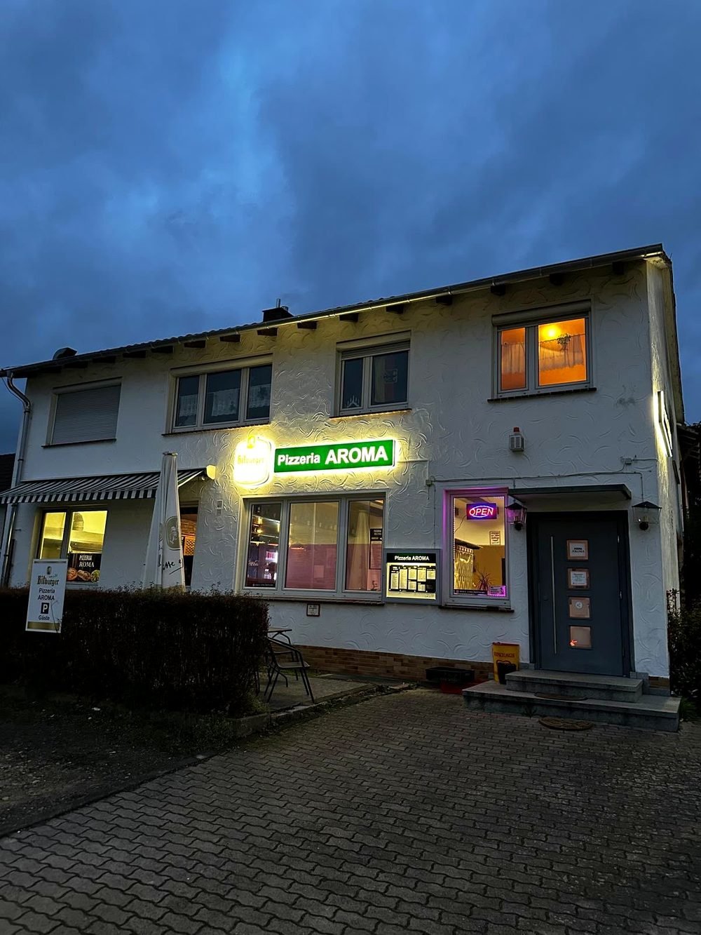 Pizzeria Aroma am Abend mit warm beleuchtetem Eingang und Außensitzplätzen